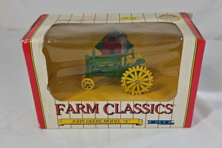 lot 74 image: Diecast John Deere Model A tractor features a green body with yellow spoked wheels and detailed engine components. Packaged in the original window box labeled Farm Classics by ERTL. Classic farm toy replica design.